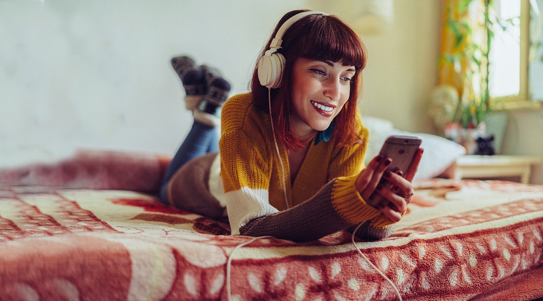 Young adult lying on her bed looking at her phone with large headphones on