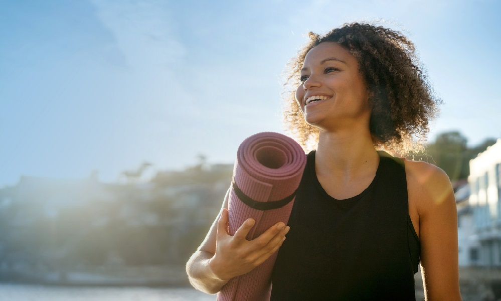 Lady with frizzy hair holding a yoga mat smiling to her left against backlit sunlight