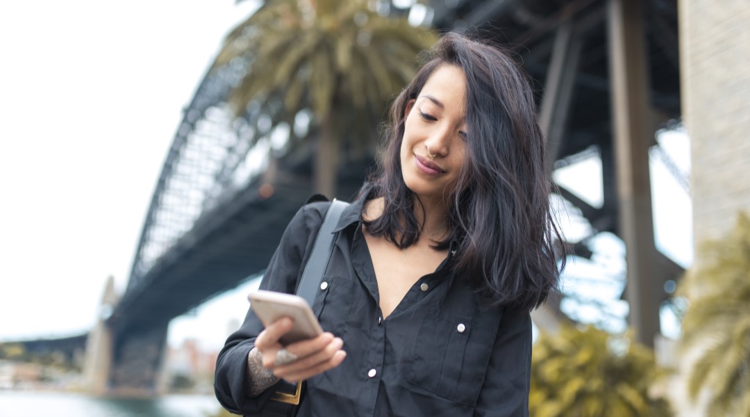 A young woman stands in front of the Sydney Harbour Bridge and uses her phone to research shares